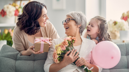 Lady giving her senior mother a present and flowers with young daughter sitting on couch